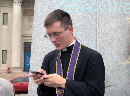 A brave priest , Fr. Stephen Schumacher, stands between angry protesters and the statue of St. Louis.